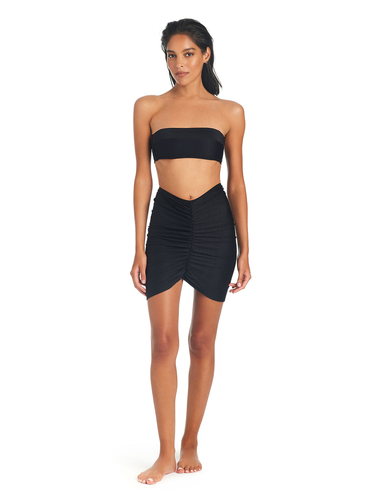 A woman with long black hair is standing barefoot on a white background, wearing the On The Water Texture Cover Up Black by Sanctuary Clothing. She faces forward with a neutral expression and has a relaxed posture. The outfit consists of a black strapless bandeau top and a high-waisted, ruched skirt that is short and asymmetrical.