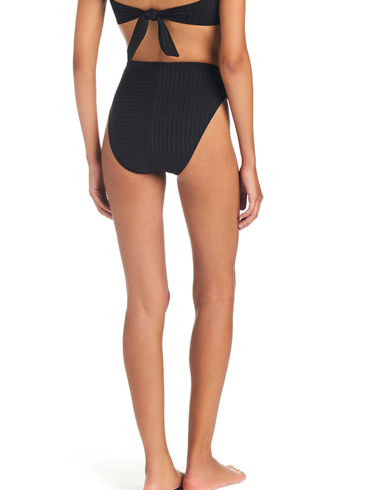 A person stands barefoot with their back facing the camera, wearing the Refresh Rib Bikini Bottom in black from Sanctuary Clothing, paired with a matching top that ties at the back. The background is plain white.