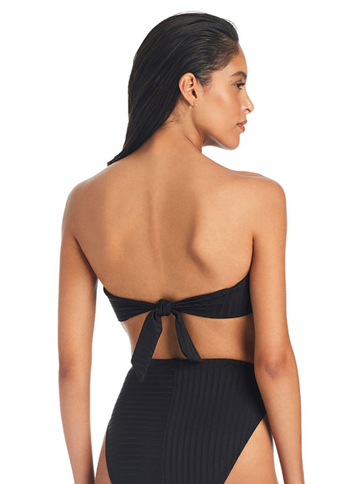 A person is shown from the back, wearing a black Refresh Rib Bandeau Bikini Top by Sanctuary Clothing, paired with high-waisted bottoms. The top is tied at the back with a knot. The person has wet, dark hair slicked back. The background is plain white.