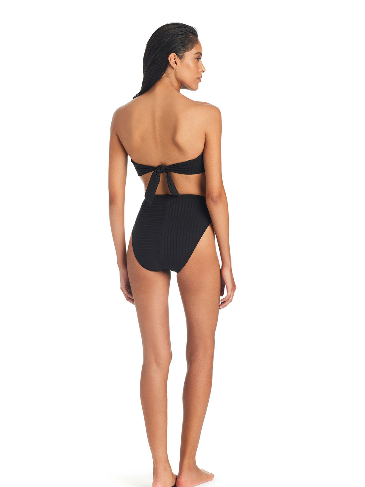 A woman stands with her back to the camera, wearing the "Refresh Rib Bikini Bottom Black" by Sanctuary Clothing, featuring a black strapless bikini top with a tied bow at the back and high-waisted bottoms. She has long, dark hair and appears to be posing against a plain, white background.