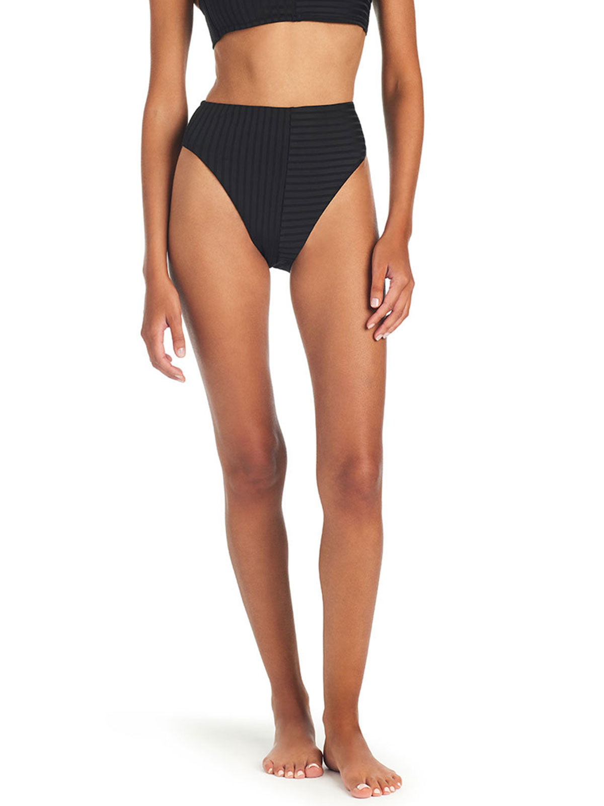 A person stands barefoot, wearing a black two-piece swimsuit from Sanctuary Clothing. The high-waisted bottom is the Refresh Rib Bikini Bottom Black, paired with a cropped top featuring wide straps. The fabric showcases a subtle textured pattern against a plain white background.