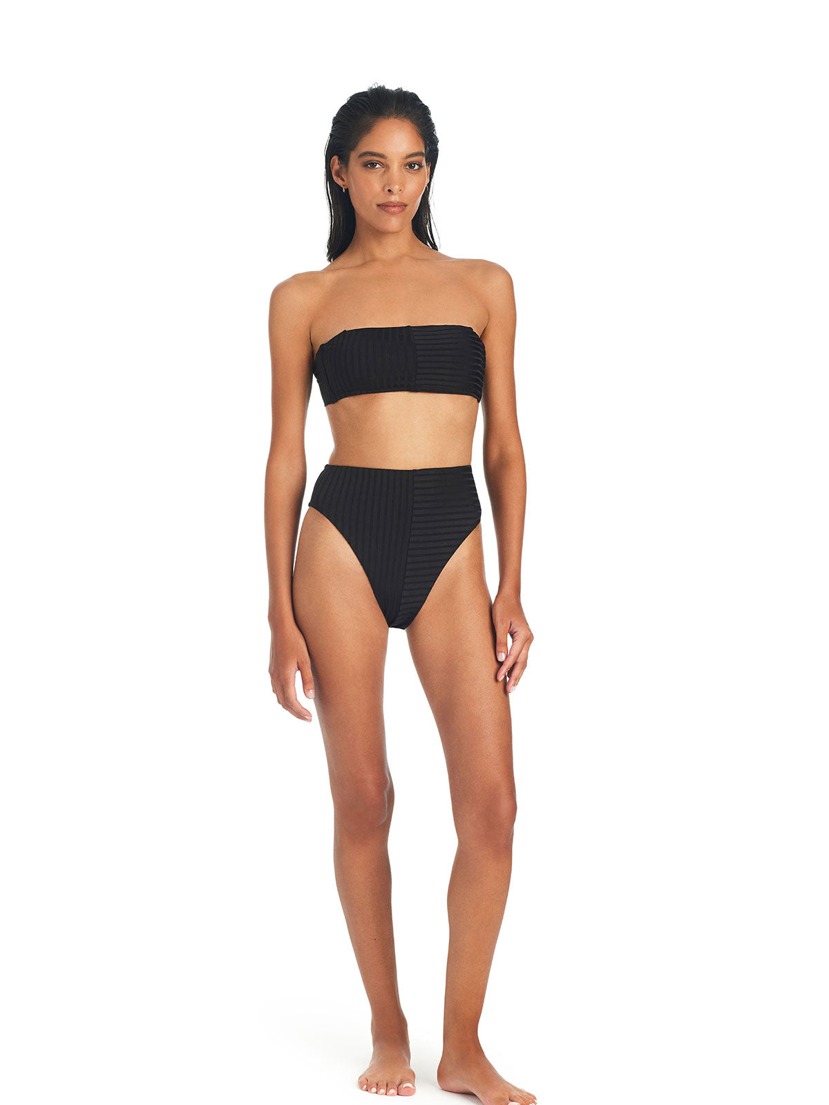 A woman with long dark hair stands barefoot against a plain white background, wearing the Sanctuary Clothing Refresh Rib Bandeau Bikini Top in black and matching high-waisted bikini bottoms. She has a neutral expression with her arms resting naturally by her sides.