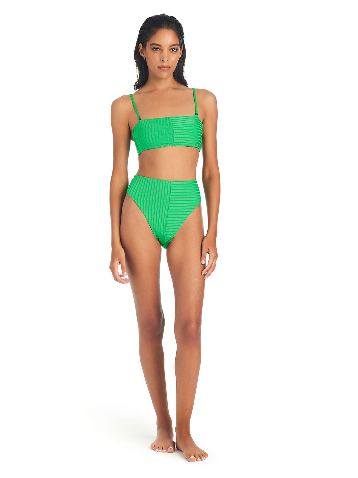 A woman is standing against a white background, wearing Sanctuary Clothing's Refresh Rib Bandeau Bikini Top in Green Flare, paired with matching high-waisted bottoms. She has long dark hair, a neutral expression, and is barefoot.