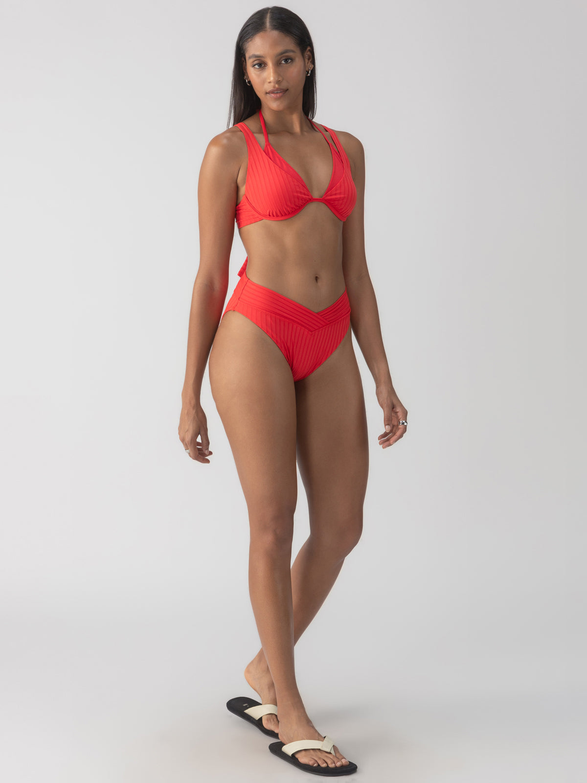 A person wearing the Sanctuary Clothing refresh rib double layer underwire bikini top in Mars Red poses against a plain background. They're standing confidently with one leg slightly forward, looking towards the camera with a slight smile, accessorized with black flip-flops.