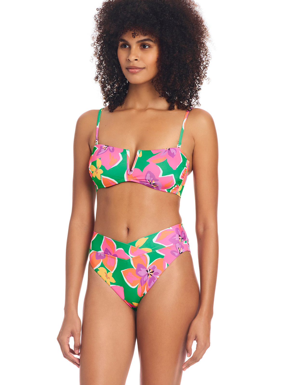 A woman with curly hair is wearing the vibrant Super Bloom V Wire Bandeau Bikini Top Multi by Sanctuary Clothing. The bikini top features a green base adorned with vibrant pink and yellow floral patterns. She stands with a relaxed posture, looking slightly to the left.