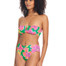 A woman is wearing a vibrant, floral-patterned bikini with a strapless top and high-waisted bottoms. The swimsuit, named the Super Bloom V Waist Bikini Bottom Multi by Sanctuary Clothing, features vivid shades of green, pink, and yellow with large flower designs. She has curly hair and is standing against a white background.