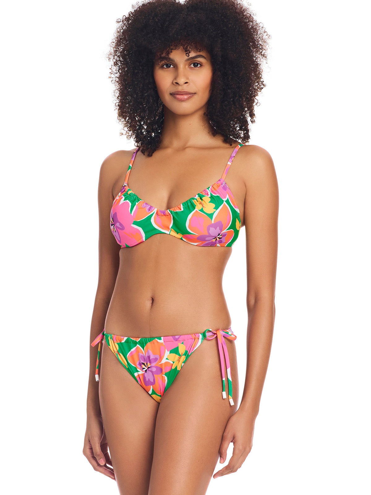 A woman with curly hair is wearing a brightly colored, floral bikini from Sanctuary Clothing. The bikini top features thin straps and a ruffled edge, while the bottom is the Super Bloom Side Tie Hipster Bikini Bottom Multi with side-tie strings. She stands against a white background, looking directly at the camera with a neutral expression.