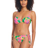 A woman with curly hair is wearing the Super Bloom Tunnel Underwire Bikini Top Multi by Sanctuary Clothing, which showcases a vibrant floral pattern in shades of pink, yellow, and green. The bikini features spaghetti straps, slight ruffles on the top edges, and tie-side bottoms. She is standing against a white background, facing forward.