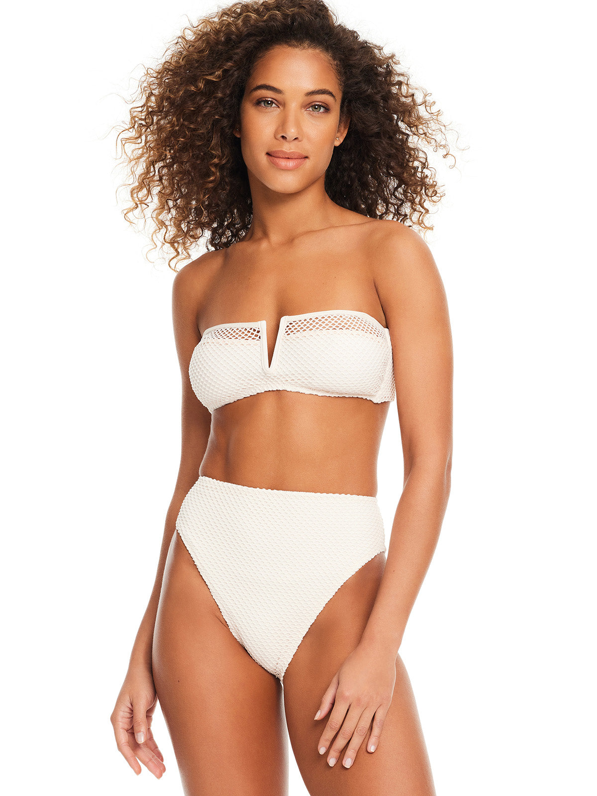 A woman with curly hair is wearing the 'Sea Crochet V Wire Bandeau Bikini Top White Sand' by Sanctuary Clothing, paired with high-waisted bikini bottoms. She is standing and smiling with her hand on her hip, against a plain white background.