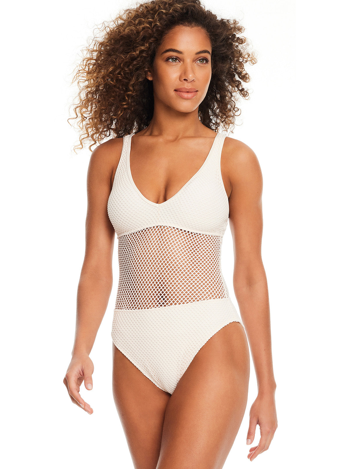 A woman with curly hair smiles while wearing the Sanctuary Clothing Sea Crochet Scoop Neck One Piece Swimsuit in White Sand, featuring a net-like, see-through midsection. She is standing against a plain white background.