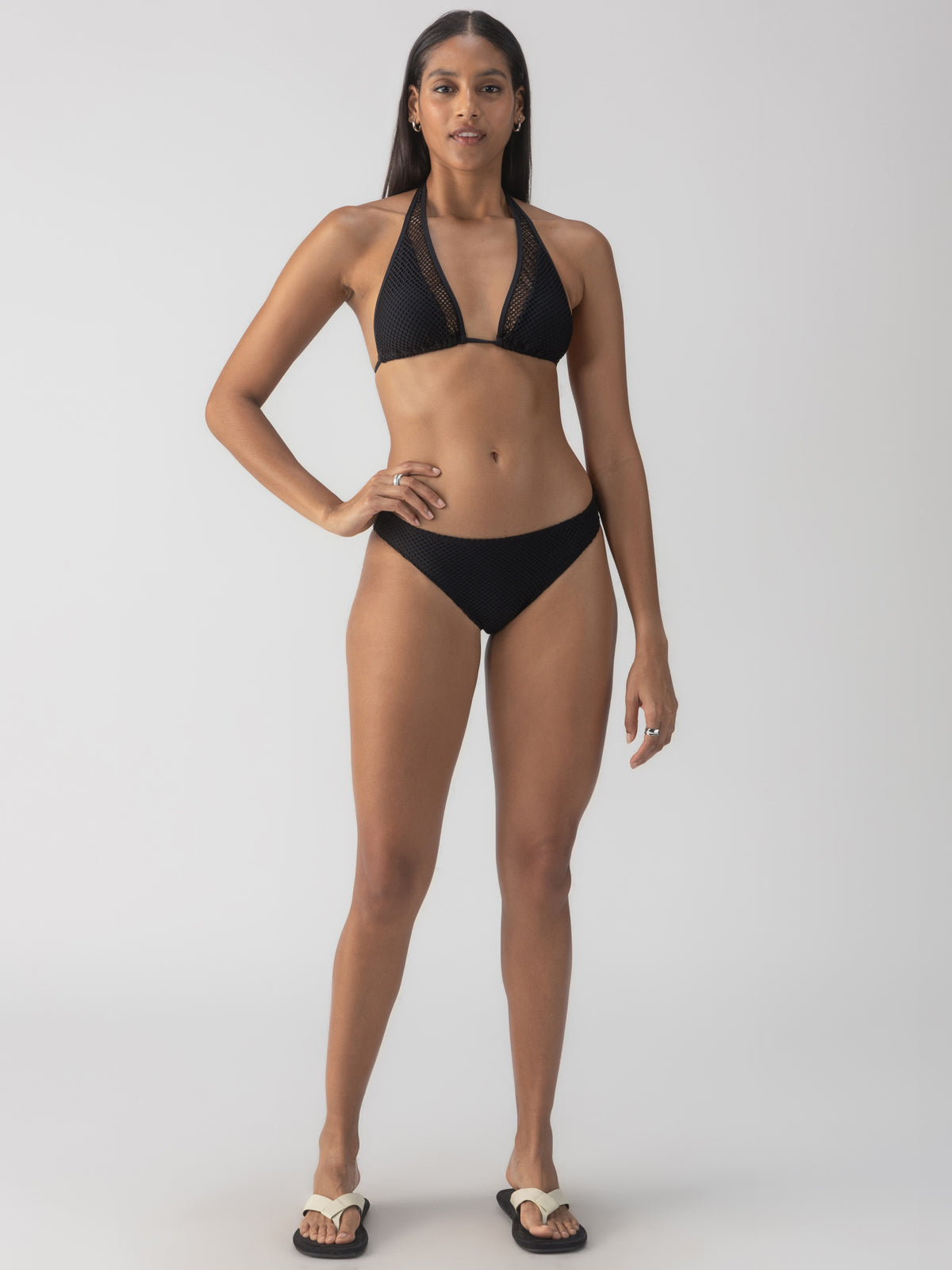 A person confidently poses against a plain background wearing the Sea Crochet Halter Tall Tri Bikini Top in black by Sanctuary Clothing, their long hair down. They stand straight with their hands by their sides and are paired with white slide sandals.