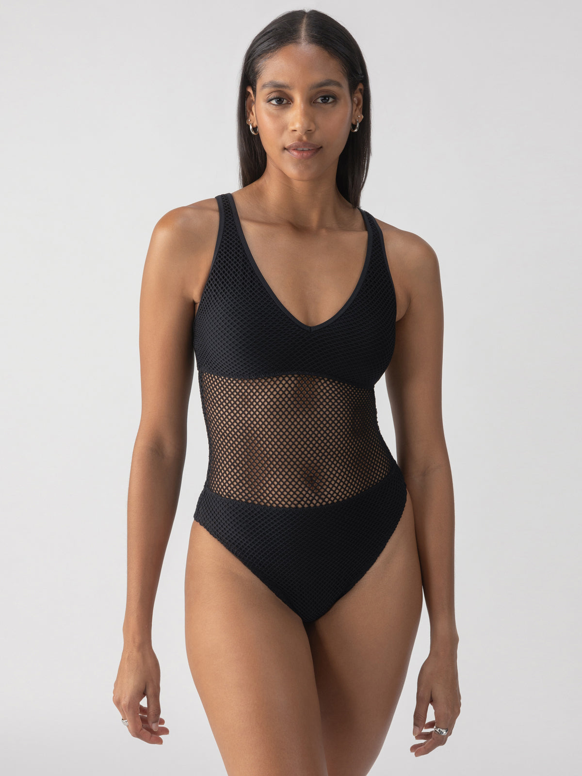 A woman wearing the Sanctuary Clothing sea crochet high leg scoop neck one piece in black stands against a neutral background. The swimsuit is designed with a scoop neckline and a sheer midsection. She has long dark hair and is adorned with minimal jewelry.