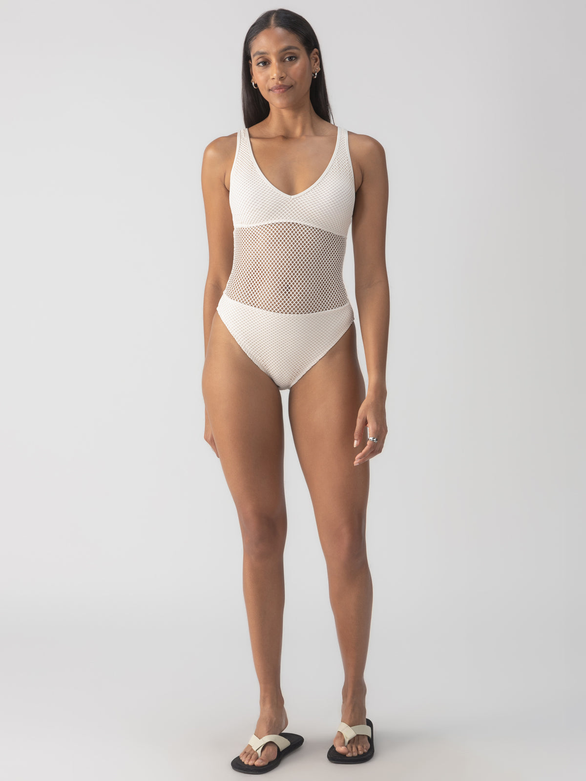 Dressed in the Sanctuary Clothing's sea crochet high leg scoop neck one piece in white sand, a person stands self-assuredly. They pair the cream-hued swimsuit, featuring a mesh panel midsection, with black and white sandals against a plain, light-colored backdrop that highlights the swimwear.