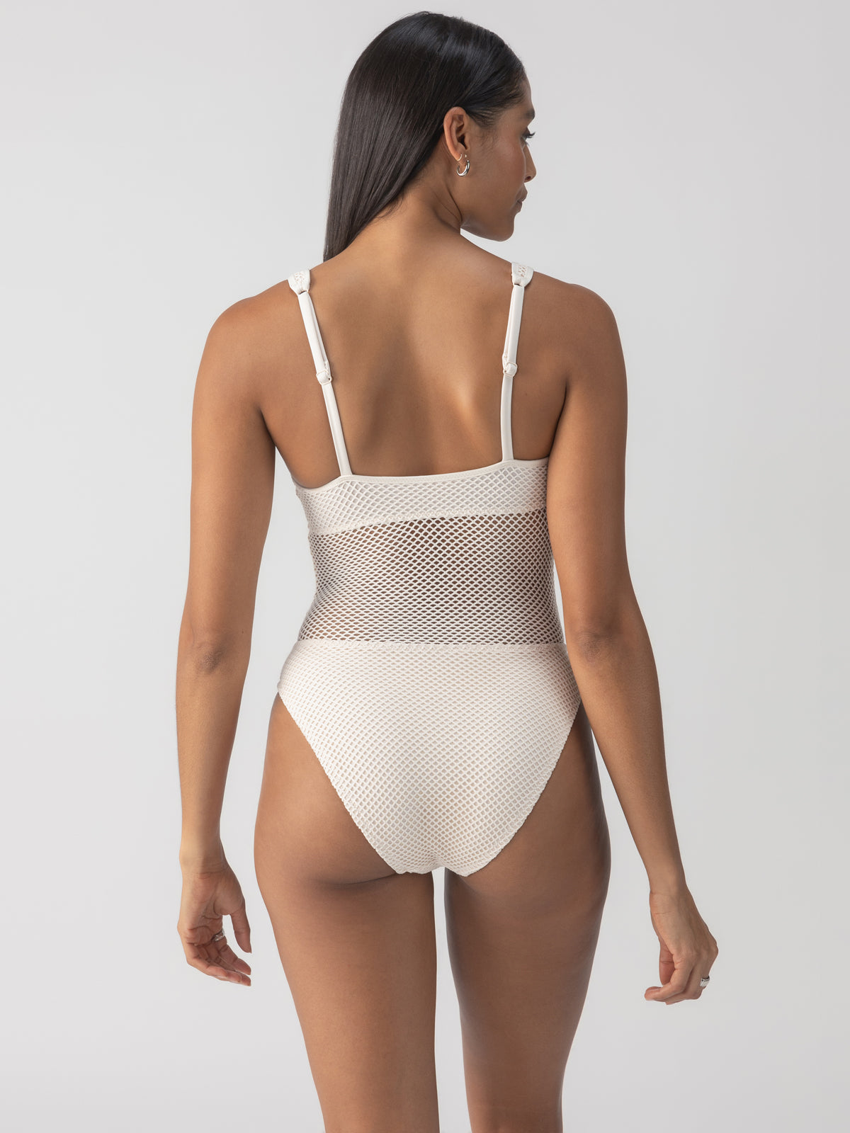 A person with long dark hair is wearing the "Sea Crochet High Leg Scoop Neck One Piece" in White Sand by Sanctuary Clothing, featuring mesh details at the waist, standing with their back to the camera against a plain light background.