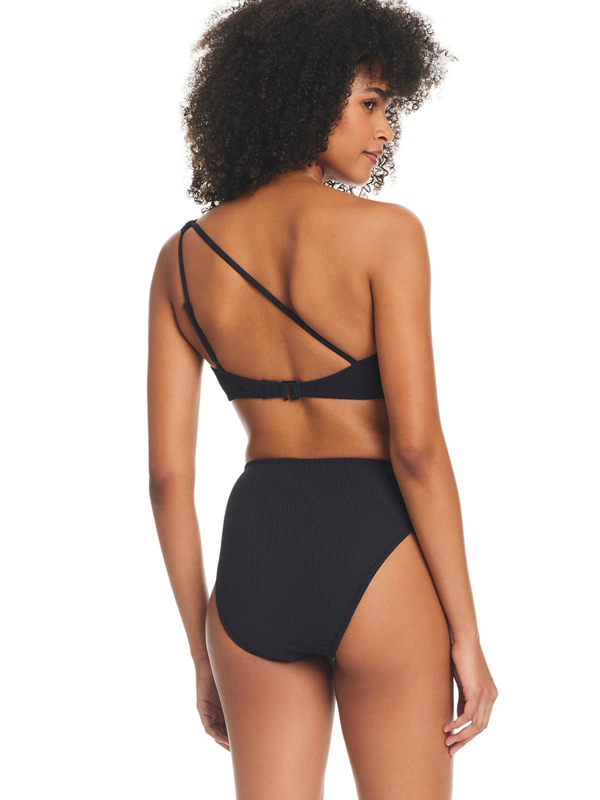 A woman with curly hair is standing and facing away from the camera, wearing the Sandbar Solids One Shoulder Bikini Top in black by Sanctuary Clothing. The high-waisted bikini features a unique crisscross strap design on the top, set against a plain white background.