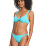 A woman with curly hair is wearing the Sanctuary Clothing Sandbar Solids Underwire Bikini Top in Curacao. The bikini top features a knot in the center, and she pairs it with simple, classic-cut bottoms. She stands facing slightly to the side, smiling softly.