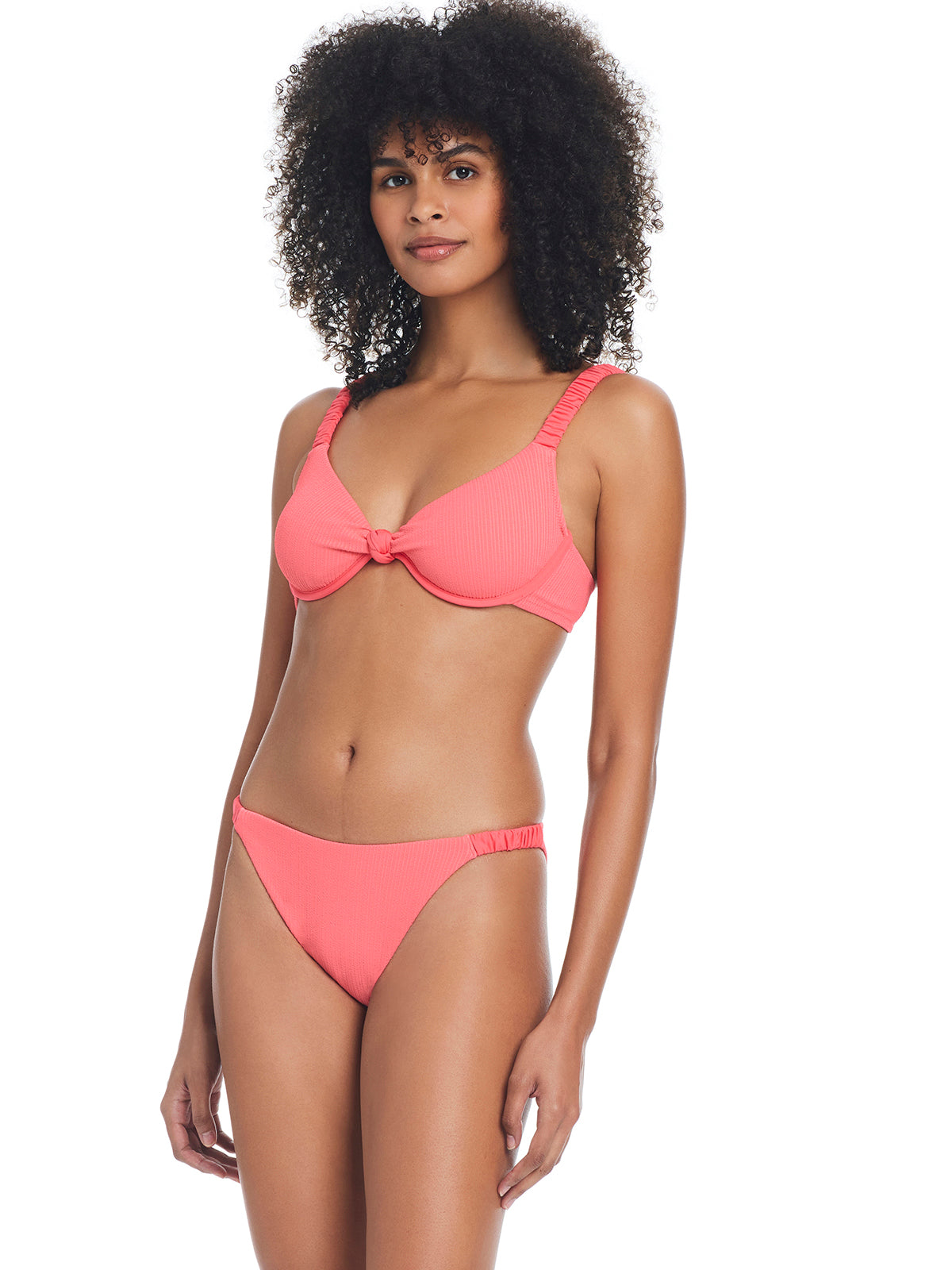 A woman with curly hair models the Sanctuary Clothing "Sandbar Solids Underwire Bikini Top" in Watermelon Sugar. The top features a knot detail in the center, and the matching bottoms have a simple, streamlined design. She stands against a plain white background, smiling slightly.