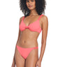 A woman with curly hair models the Sanctuary Clothing "Sandbar Solids Underwire Bikini Top" in Watermelon Sugar. The top features a knot detail in the center, and the matching bottoms have a simple, streamlined design. She stands against a plain white background, smiling slightly.