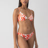 A person with long hair is wearing the Sanctuary Clothing "Sol Searcher OTS Underwire Bikini Top" in white sand, featuring an orange and white swirl pattern. They are standing in front of a plain gray background, maintaining a neutral expression while looking toward the camera.