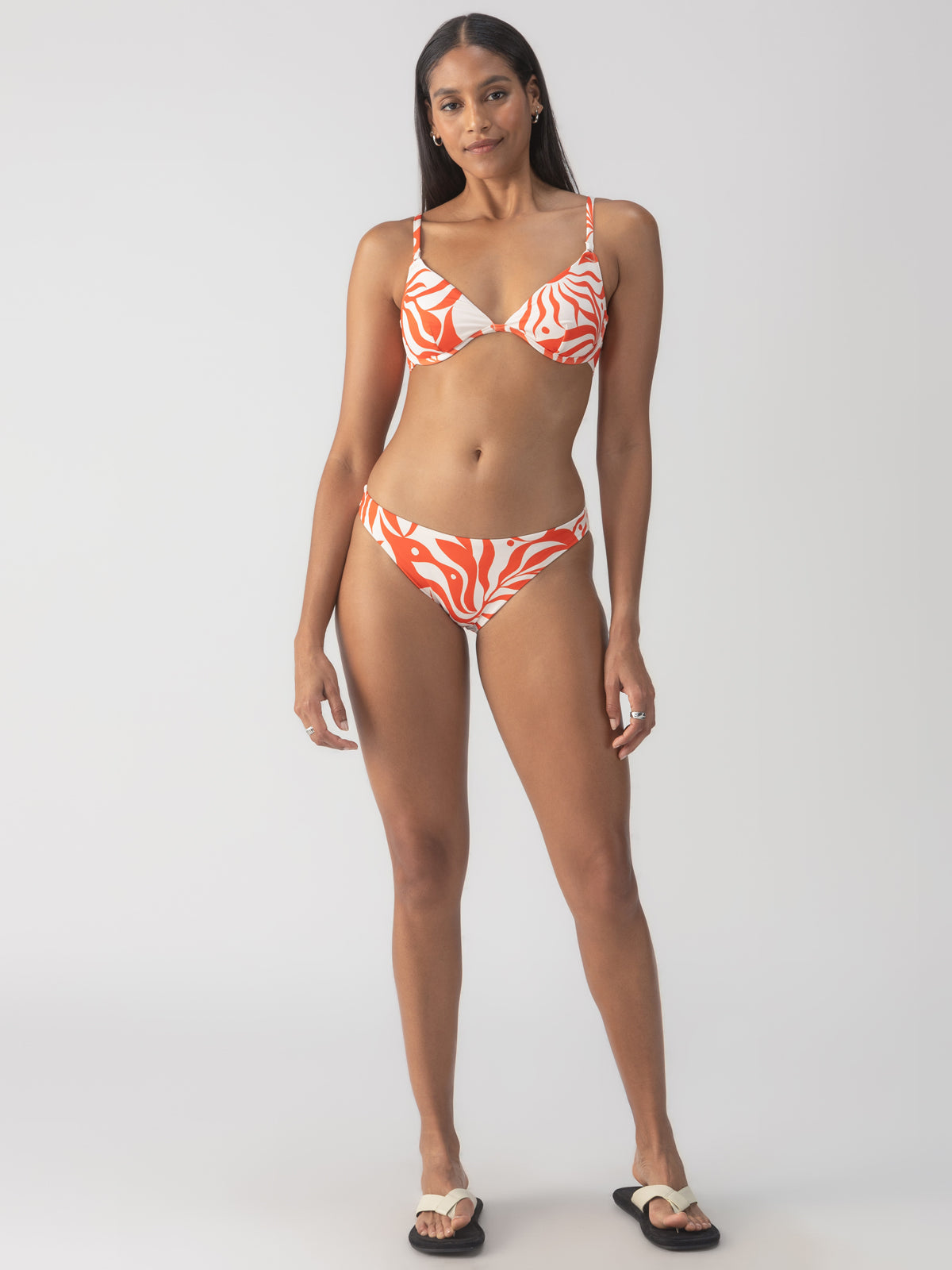 A woman stands confidently wearing the "Sol Searcher Cinch Back Hipster Bikini Bottom" in White Sand from Sanctuary Clothing. Her long hair is down, and she's wearing black flip-flops. The plain background highlights her stylish swimwear.