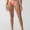 A person stands against a plain backdrop wearing the vibrant "sol searcher cinch back hipster bikini bottom" in white sand by Sanctuary Clothing. They are also sporting black and white sandals. The image captures them from the shoulders down.