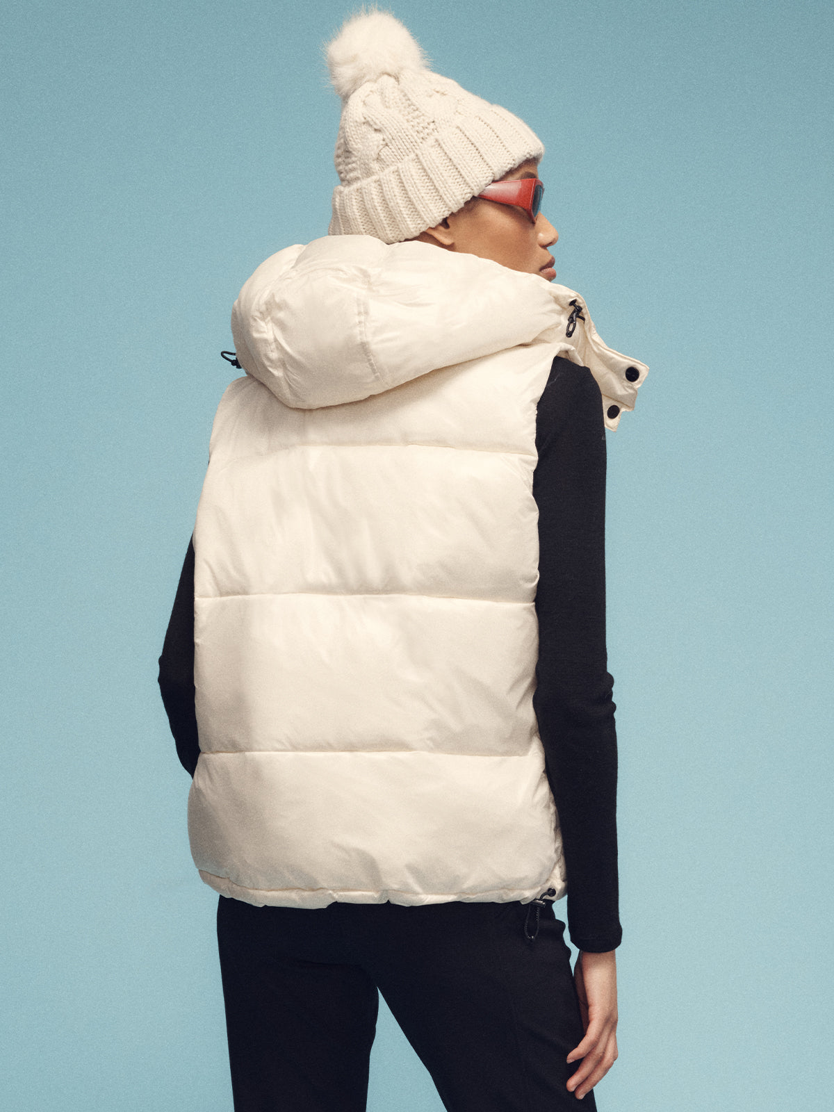 A person facing away from the camera is sporting a Sanctuary Clothing puffer up vest in sugar cookie color and wearing a knitted beanie with a pom-pom. They have sunglasses on and are dressed in a black long-sleeve shirt, set against a light blue background.