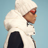 A person in profile is wearing the Sanctuary Clothing puffer up vest sugar cookie over a black shirt, paired with a white knitted beanie featuring a pom-pom and red sunglasses, all set against a light blue background.