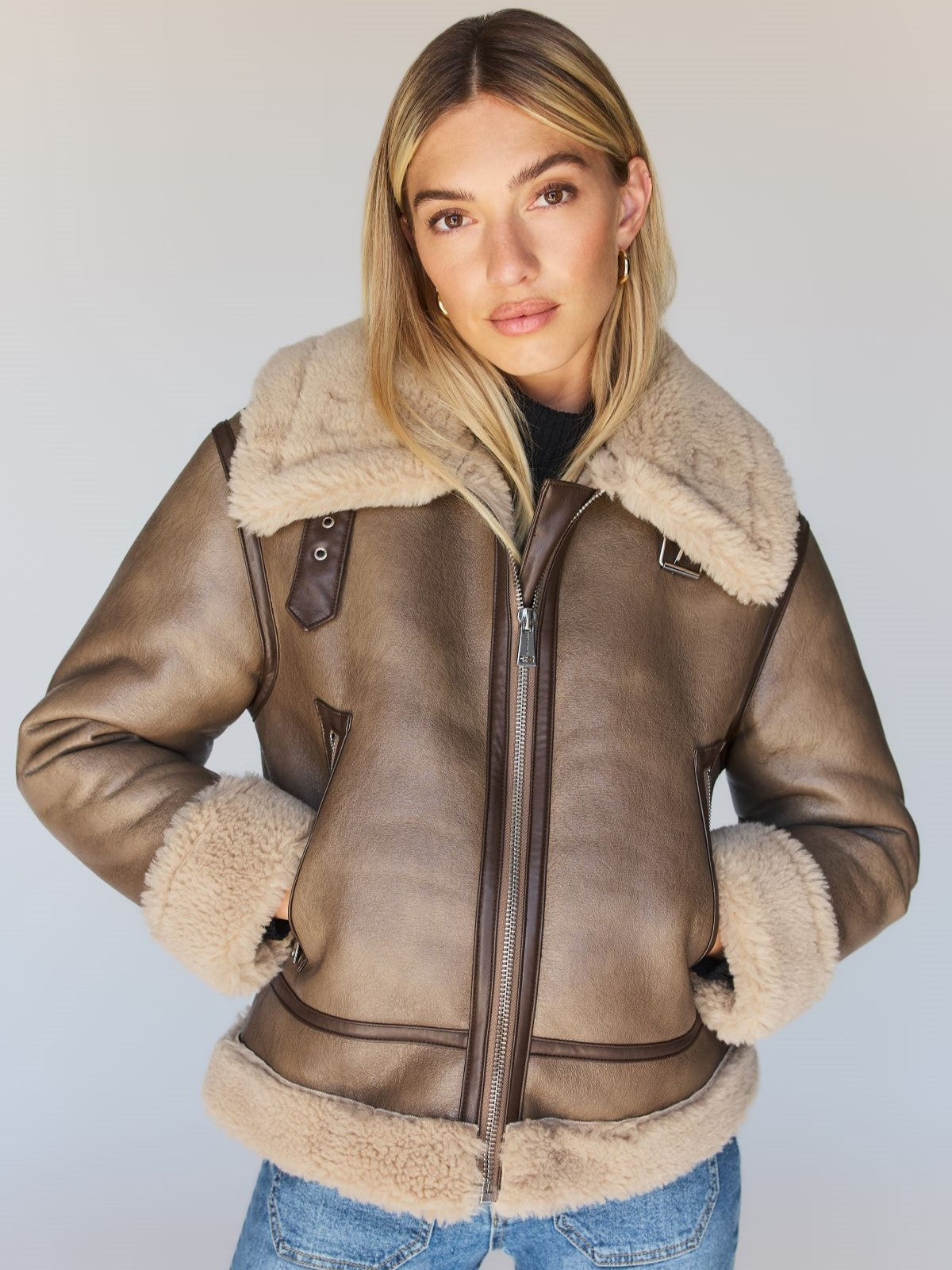 A person with long blonde hair wears the Zarina Shearling Coat Mink Beige by Sanctuary Clothing, featuring a zipper and plush fur collar and cuffs. They stand with hands in pockets against a neutral background.