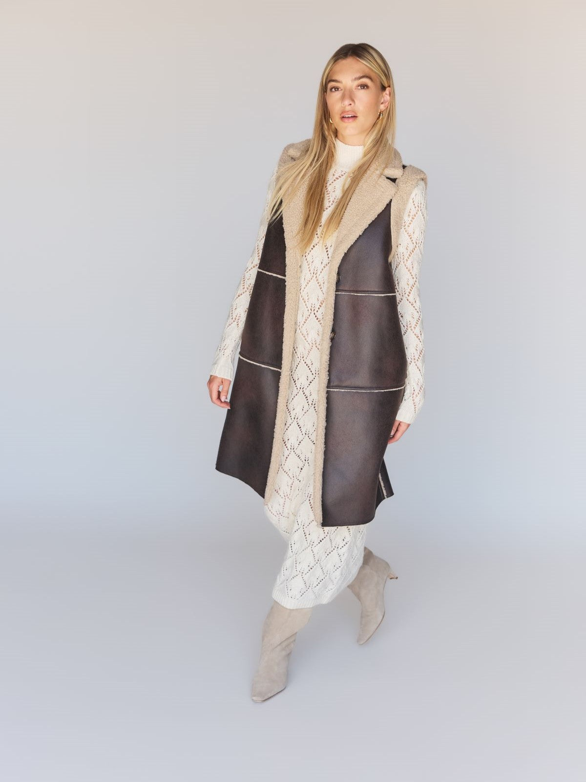 A woman wearing the Espresso Paisley Shearling Vest by Sanctuary Clothing over a white, patterned dress with beige boots, posing against a neutral background.