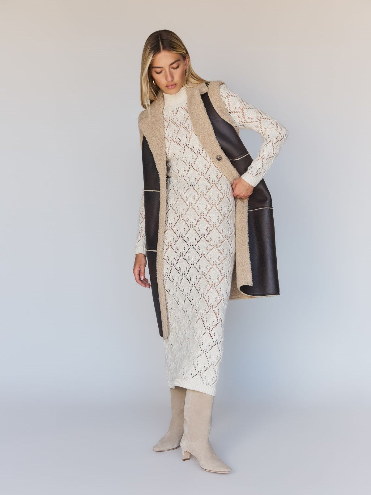 A woman is wearing a long, cream-colored, patterned dress paired with Sanctuary Clothing's Paisley Shearling Vest in Espresso. She also has on light beige knee-high boots and stands against a neutral background. Her hair is shoulder-length and blonde.