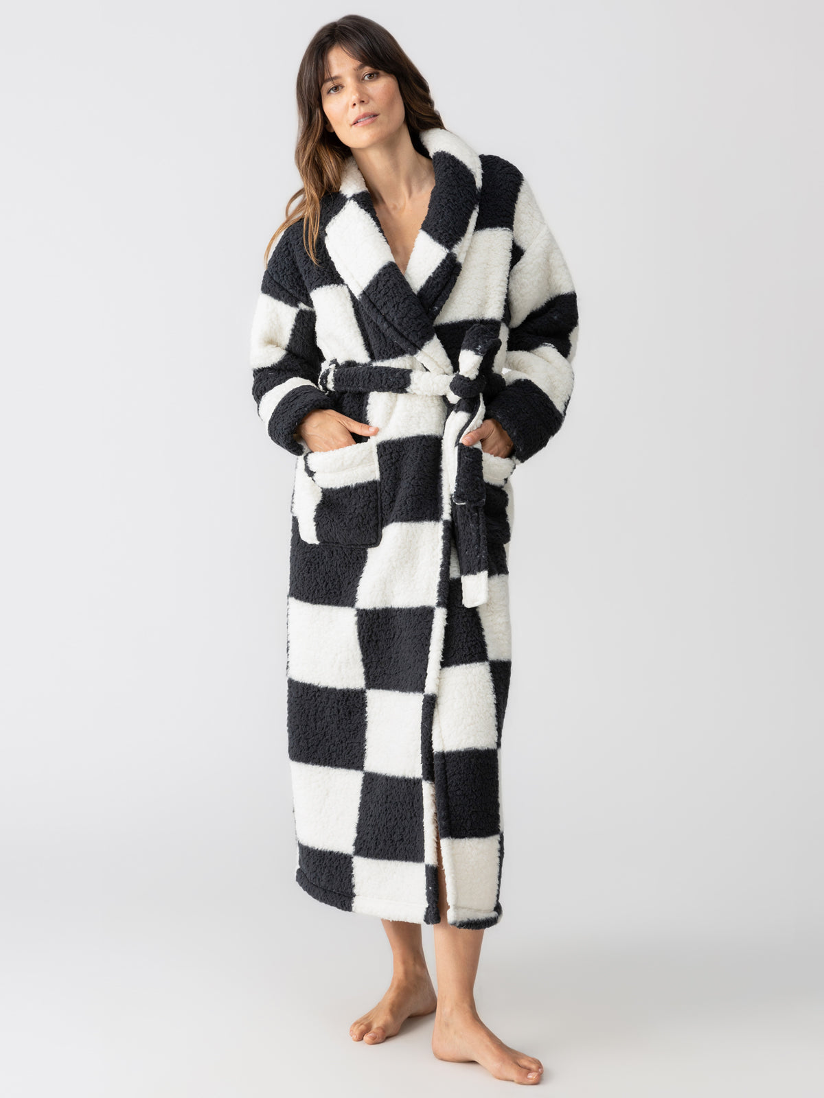 A person models the Sanctuary Clothing shawl collar teddy robe in a black checkered pattern, featuring convenient pockets and a coordinating belt. They are barefoot against a simple, light-colored backdrop.