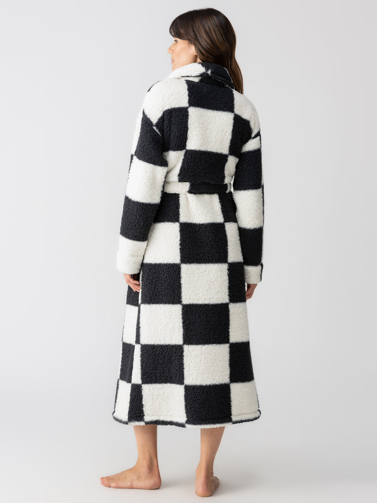 A person wearing the Sanctuary Clothing's shawl collar teddy robe black check, standing barefoot on a plain white background, facing away from the camera. The robe features a belted waist and is made from a cozy, textured material.