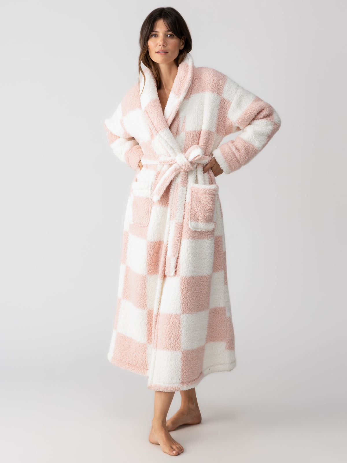 A person wearing the Sanctuary Clothing shawl collar teddy robe in rose check stands barefoot against a plain background. This plush bathrobe features large pockets and a belt, and the person has their hands on their hips.