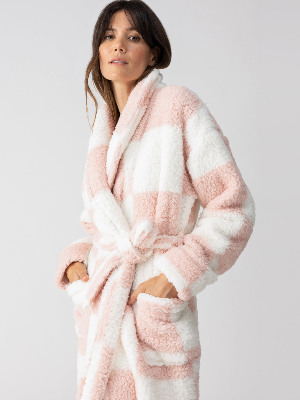 A person is wearing the Shawl Collar Teddy Robe in Rose Check by Sanctuary Clothing. They have their hands in the robe's pockets and are looking forward with a slight smile against a plain, light-colored background.