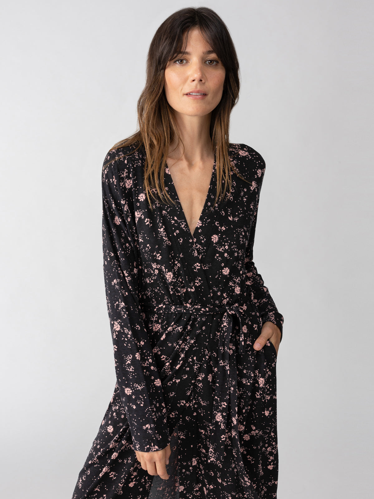 A person with long brown hair stands against a plain background, wearing the Maxi Cardi Falling Flowers dress by Sanctuary Clothing. The dress, in black with pink floral prints, features long sleeves and a V-neckline and is tied at the waist. The individual has a neutral expression and looks directly at the camera.