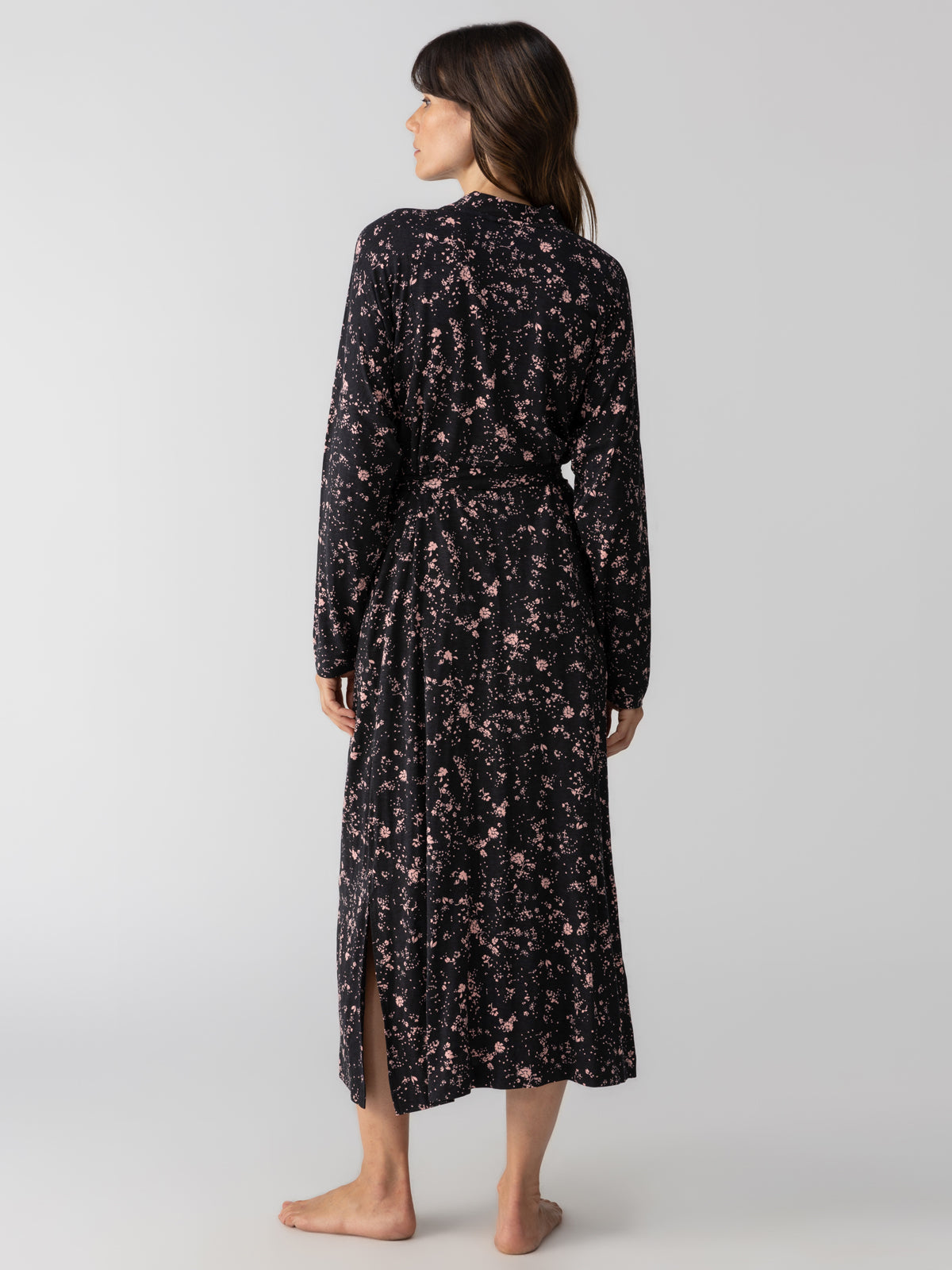 A woman stands barefoot, facing away from the camera. She is wearing a Maxi Cardi Falling Flowers by Sanctuary Clothing, featuring a long black dress with pink floral patterns, long sleeves, and a waist tie. She has long brown hair and is set against a plain, light gray background.