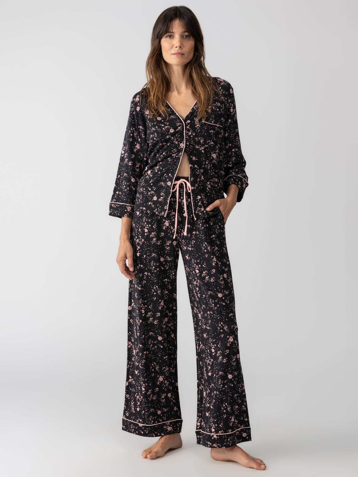 A woman stands barefoot against a plain background, wearing the Lounge Set Falling Flowers from Sanctuary Clothing, a matching black pajama set adorned with a floral pattern. The top features long sleeves and is partially unbuttoned, while the pants include an adjustable drawstring waistband. She has a relaxed expression.