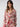 A person with long hair is wearing the Sanctuary Clothing's "Notch and Pant Set Winter Rose" pajamas adorned with a pink and red rose pattern. The top features a pocket and red piping, while their hand rests casually in the pants pocket as they stand against a plain background.