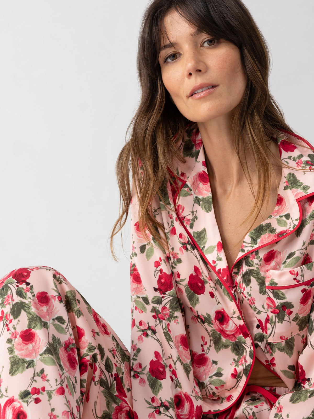 A woman with long brown hair is comfortably seated against a light background, dressed in the "Notch and Pant Set Winter Rose" by Sanctuary Clothing. The pajama set showcases red and green rose patterns, complementing her soft and serene expression.