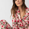 A woman with long brown hair is comfortably seated against a light background, dressed in the "Notch and Pant Set Winter Rose" by Sanctuary Clothing. The pajama set showcases red and green rose patterns, complementing her soft and serene expression.