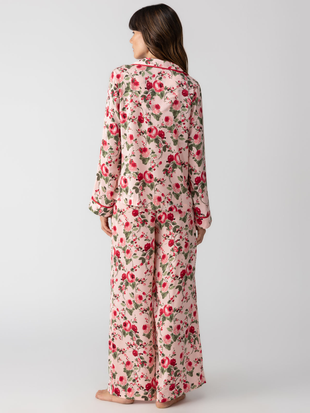 A person stands facing away, wearing the Sanctuary Clothing "notch and pant set winter rose," a matching floral print pajama ensemble with long sleeves and wide-legged pants. The background is plain and light-colored.