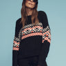 A woman in Sanctuary Clothing's intarsia sweater black multi and a hat poses against a blue background. She wears dark sunglasses and has long hair. The sweater features a geometric design in red, white, and black colors.