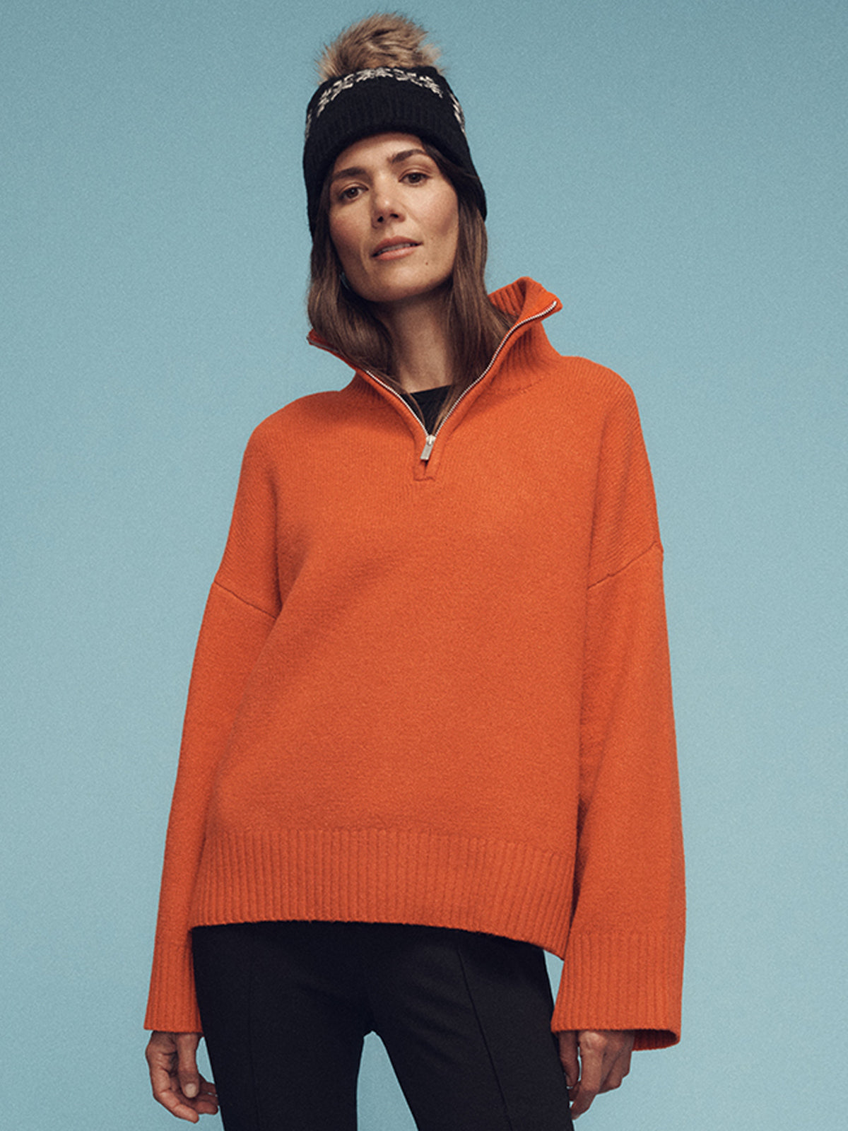 A person wearing Sanctuary Clothing's 1/4 zip cashmere-like sweater in ski orange and a black beanie with a pom-pom stands against a blue background, looking confidently into the camera. The sweater has a high collar with a half-zip.