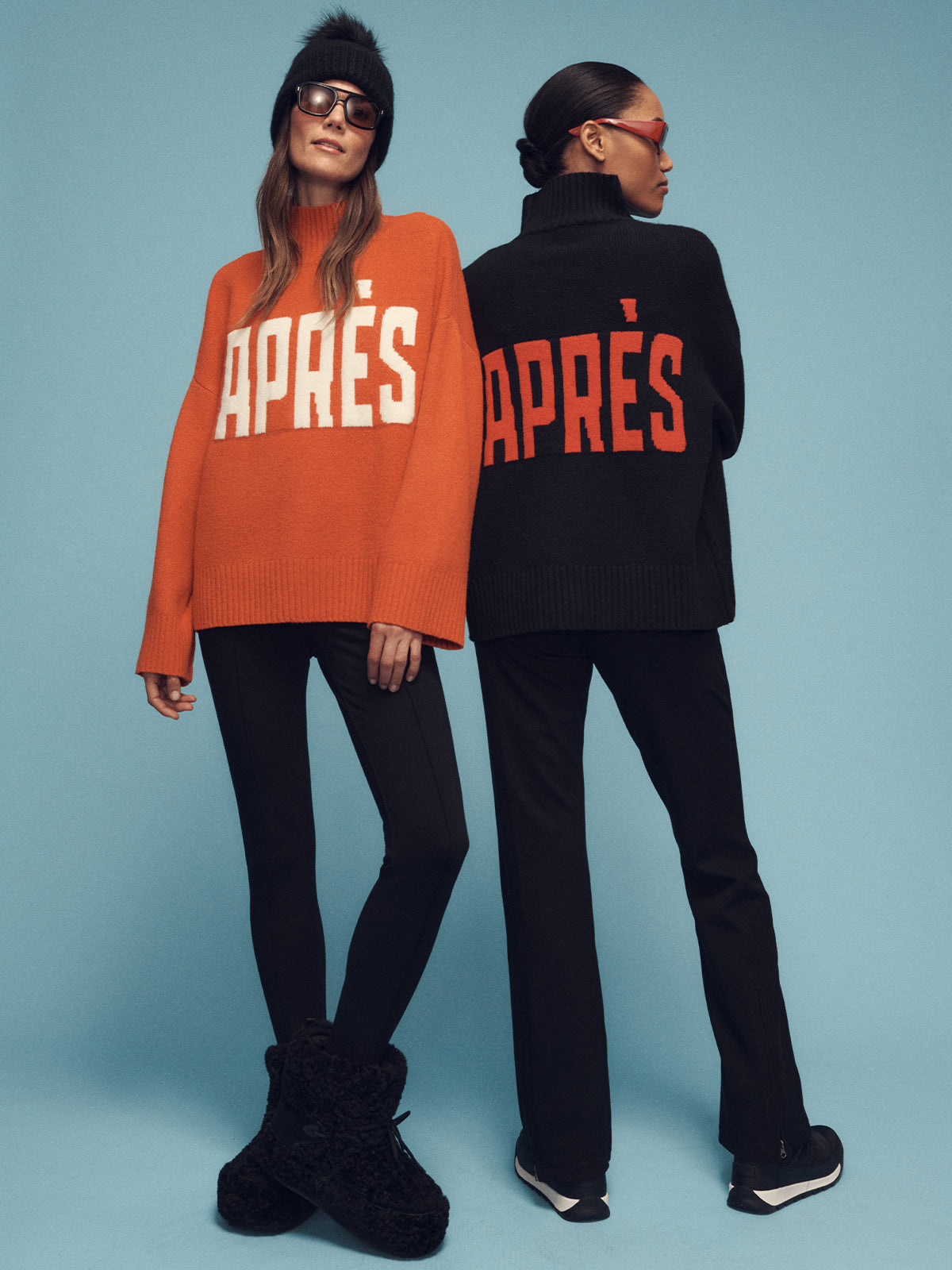 Two models pose against a blue background wearing oversized Sanctuary Clothing sweaters with "APRÈS" in bold letters. The model on the left wears a mars red sweater, and the one on the right wears a black sweater. Both are styled with black pants and stylish sunglasses.