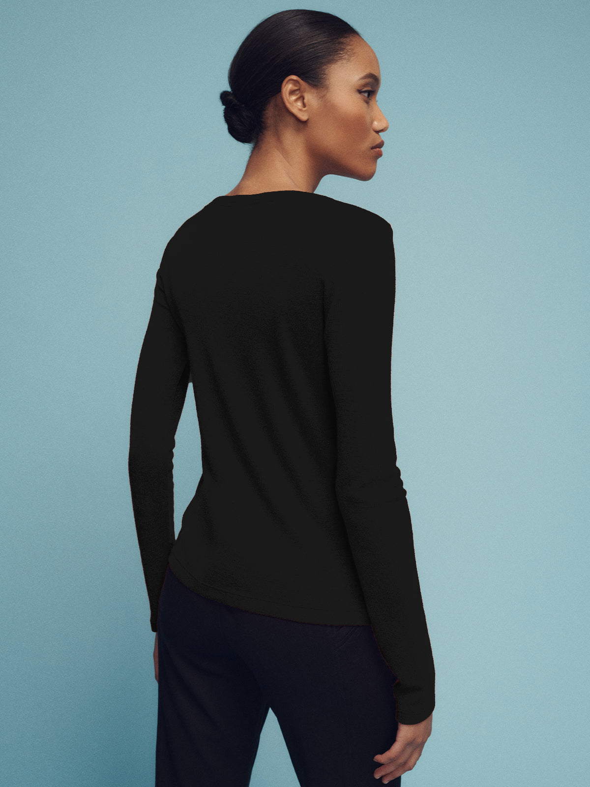 A person with dark hair tied back is wearing the Sanctuary Clothing long sleeve wool perfect tee in black, along with dark pants, standing against a light blue background, facing away.