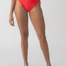 A person is wearing the Sanctuary Clothing refresh rib V-waist high-leg bikini bottom in Mars Red, accompanied by black sandals with white straps. The background is plain and minimalistic, highlighting the swimwear and sandals.