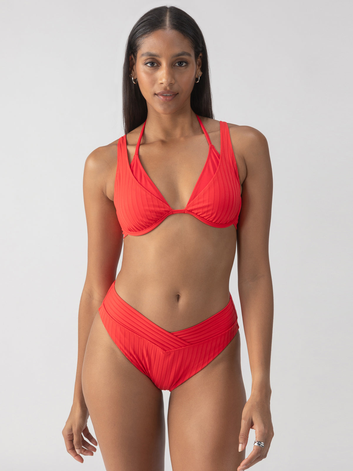 A person stands confidently, showcasing the vibrant refresh rib v waist high leg bikini bottom in mars red by Sanctuary Clothing. The backdrop is a simple light grey, accentuating the striking swimwear and the model's composed expression.