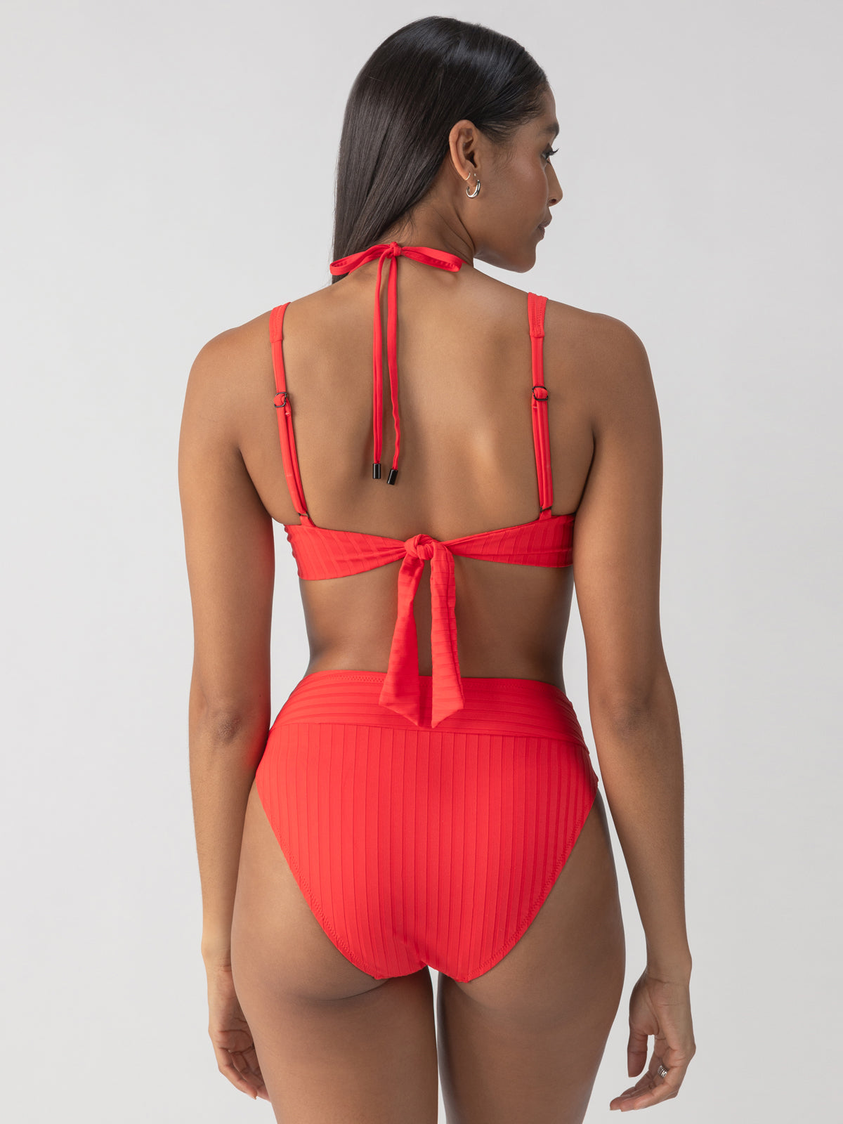 A person stands facing away, wearing the Refresh Rib V Waist High Leg Bikini Bottom in Mars Red from Sanctuary Clothing. The bikini features a halter neck tie and a subtle ribbed texture. The plain, light background accentuates the vibrant swimwear.