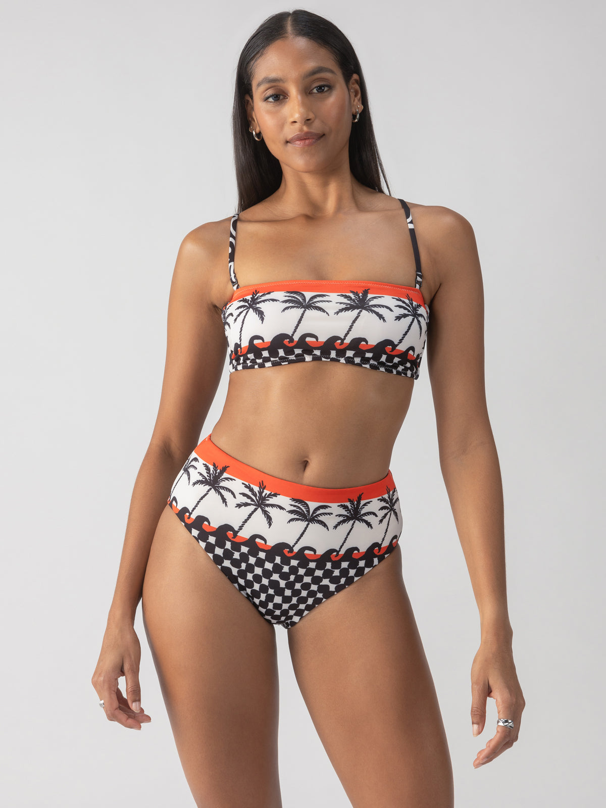 A person wearing a stylish bikini featuring palm tree and checker designs with vibrant orange accents. The top is a bandeau style, and the swimsuit includes the "Beach Con Reversible High Rise Bikini Bottom" in black by Sanctuary Clothing. The background is a light gray.