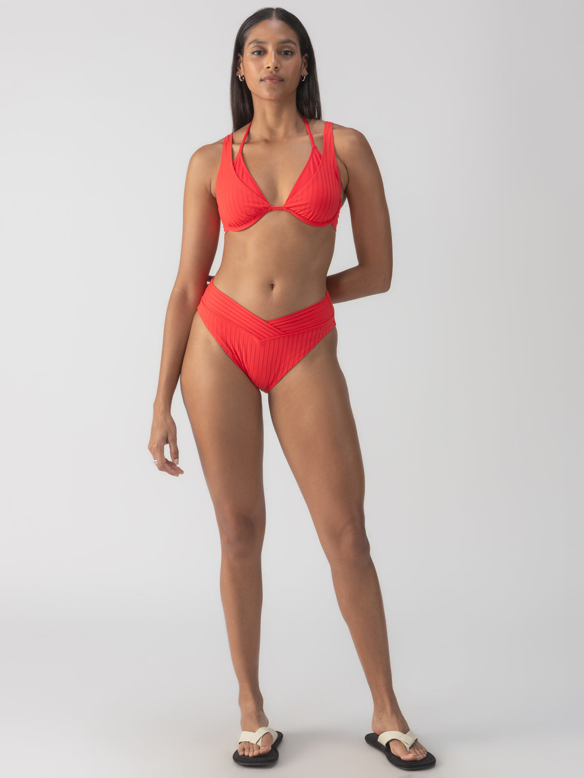 A person wearing the Sanctuary Clothing refresh rib v waist high leg bikini bottom in mars red poses against a neutral gray background. They are also wearing black and white sandals, standing confidently while looking at the camera.